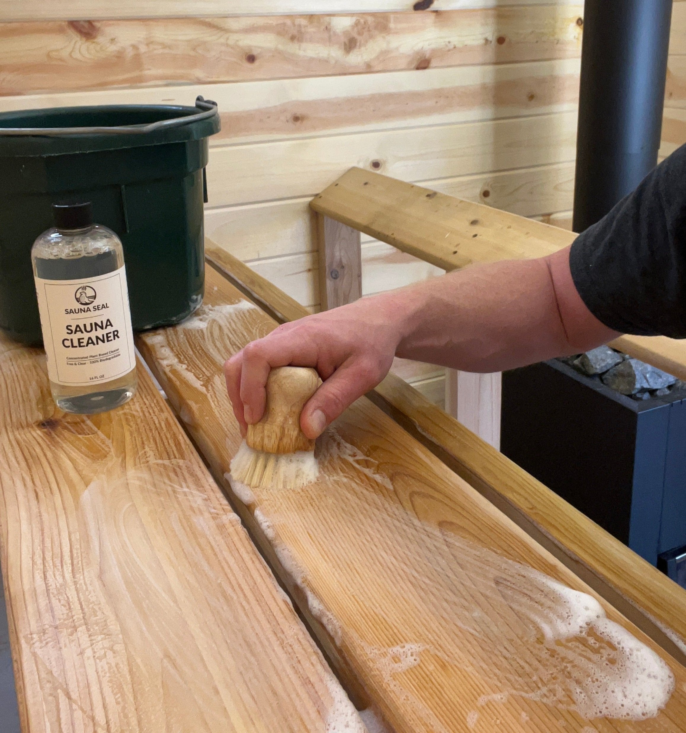 Person cleaning a wooden sauna surface with a brush and a bottle of sauna cleaner.