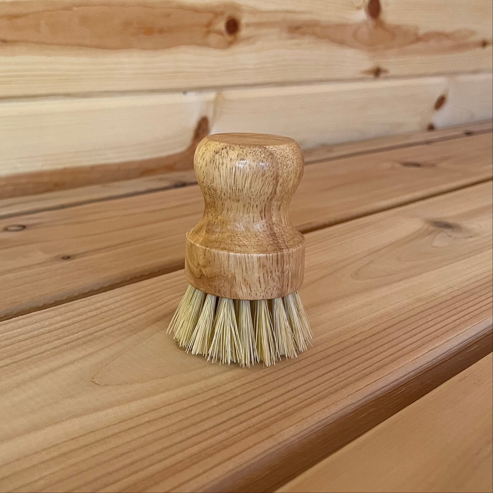 A scrub brush with a wooden handle and natural fiber bristles, placed on a wooden surface.